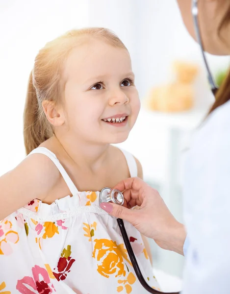 Médico examinando a un niño por estetoscopio en una clínica soleada. Feliz niña sonriente paciente vestida con vestido de color brillante es en la inspección médica habitual — Foto de Stock