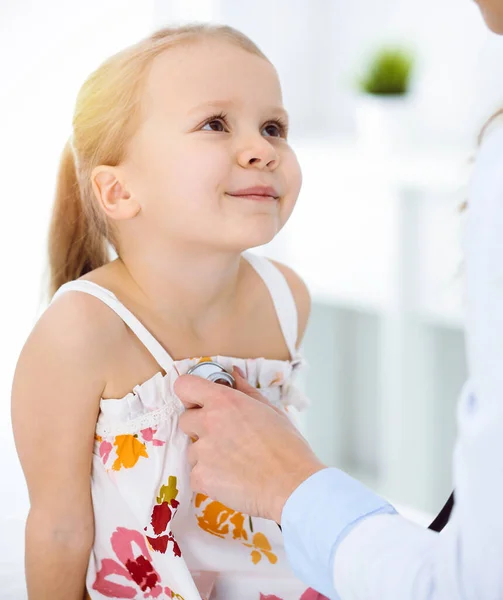 Doutor examinando uma criança por estetoscópio na clínica ensolarada. Feliz sorrindo paciente menina vestida com vestido de cor brilhante é na inspeção médica habitual — Fotografia de Stock