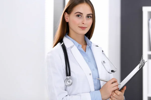 Läkarkvinnan fyller i läkarformulär när hon står nära fönstret på sjukhusets kontor. Glad läkare på jobbet. Medicin och hälsovård — Stockfoto