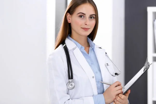 Medico donna riempiendo modulo medico mentre in piedi vicino alla finestra in ufficio ospedaliero. Medico felice al lavoro. Medicina e concetto di assistenza sanitaria — Foto Stock