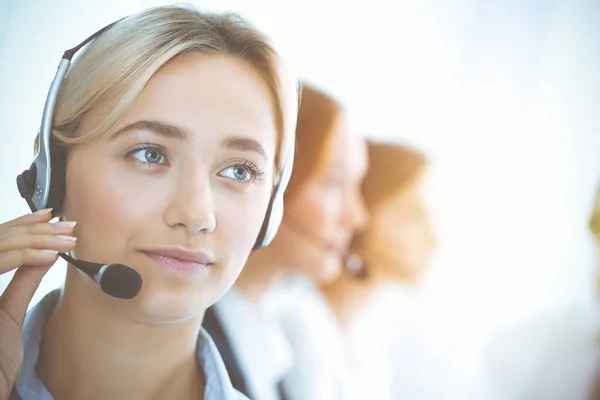 Femme d'affaires souriante joyeuse avec écouteurs consultation des clients. Groupe de différents opérateurs téléphoniques au travail dans un bureau ensoleillé.Concept de centre d'appels et de gens d'affaires — Photo