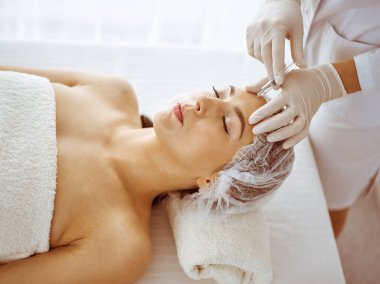 Güzellik uzmanı, genç esmer kadının yüzüne şırıngayla güzellik prosedürü uyguluyor. Kozmetik ilaç ve ameliyat, güzellik iğneleri.