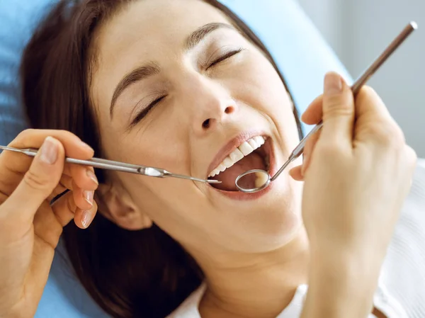Leende brunett kvinna som undersöks av tandläkare på tandkliniken. Händerna på en läkare som håller dentala instrument nära patientens mun. Friska tänder och medicin koncept — Stockfoto