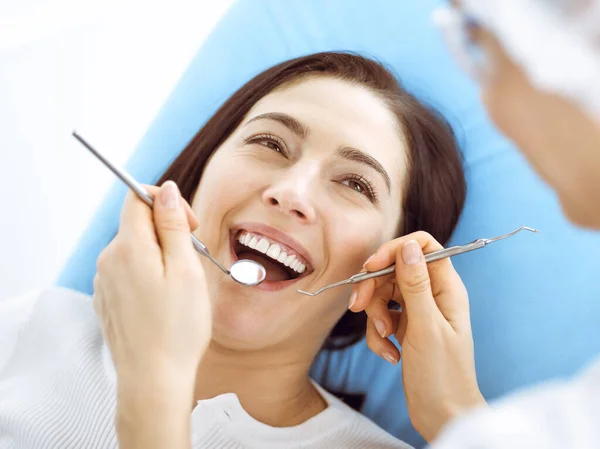 Uśmiechnięta brunetka badana przez dentystę w klinice dentystycznej. Ręce lekarza trzymającego instrumenty stomatologiczne w pobliżu ust pacjenta. Zdrowe zęby i koncepcja medycyny — Zdjęcie stockowe