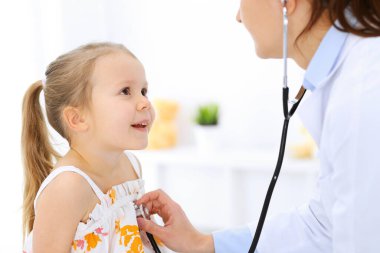 Doktor steteskopla küçük bir kızı muayene ediyor. Her zamanki tıbbi muayenede gülümseyen çocuk hasta. Tıp ve sağlık hizmetleri kavramları