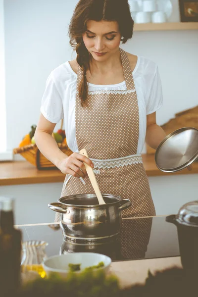 젊은 갈색 머리 여자가 부엌에서 수프를 만들고 있어. 손에 나무 숟가락을 들고 있는 주부. 식품과 건강 개념 — 스톡 사진