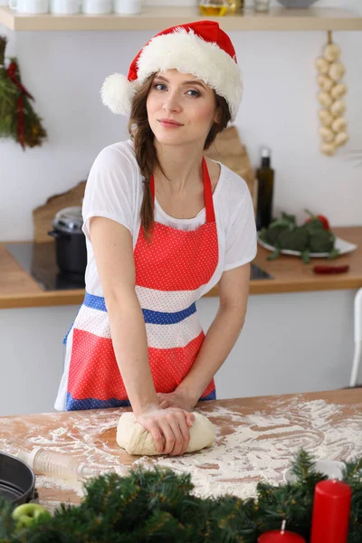 Νεαρή μελαχρινή γυναίκα μαγειρεύει πίτσα ή χειροποίητα ζυμαρικά φορώντας το καπέλο του Αϊ Βασίλη στην κουζίνα. Νοικοκυρά που ετοιμάζει ζύμη σε ξύλινο τραπέζι — Φωτογραφία Αρχείου