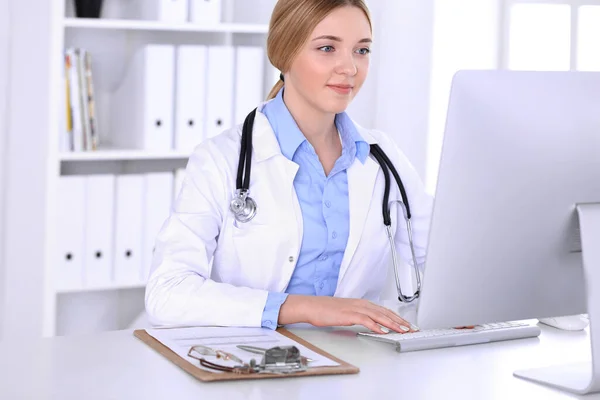 Mujer joven médico en el trabajo en el hospital mirando monitor de escritorio PC. El médico controla los registros de la historia de la medicación y los resultados del examen. Concepto de medicina y salud — Foto de Stock