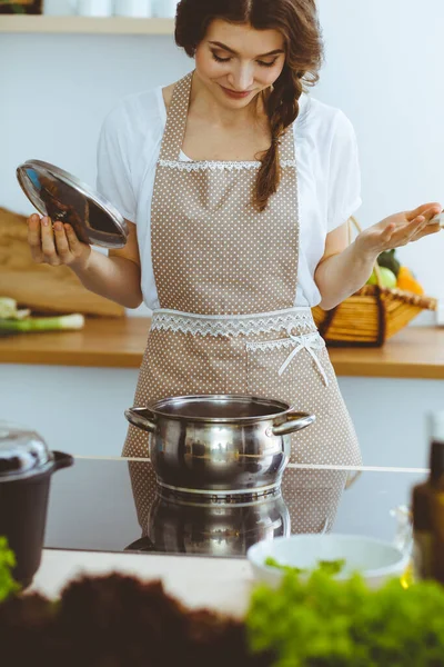 젊은 갈색 머리 여자가 부엌에서 수프를 만들고 있어. 손에 나무 숟가락을 들고 있는 주부. 식품과 건강 개념 — 스톡 사진