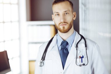 Kızıl sakallı doktor çalışma yerinin yanındaki güneşli klinikte dikiliyor. Hekim portresi. Tıp konsepti