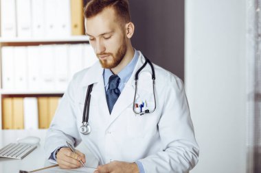 Dost canlısı kızıl sakallı doktor güneşli klinikteki panoda oturuyor ve yazıyor. Tıp konsepti