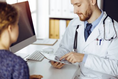 Dost canlısı kızıl sakallı doktor ve hasta kadın klinikte oturup tablet bilgisayar kullanırken sağlık muayenesini tartışıyorlar. Tıp konsepti