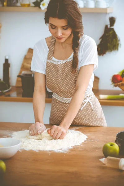 Νεαρή μελαχρινή γυναίκα μαγειρεύει πίτσα ή χειροποίητα ζυμαρικά στην κουζίνα. Νοικοκυρά ετοιμάζει ζύμη σε ξύλινο τραπέζι. Έννοια της διατροφής, των τροφίμων και της υγείας — Φωτογραφία Αρχείου