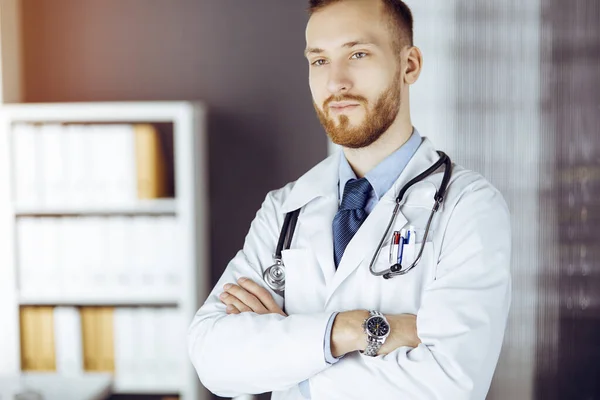 Red-bearded doctor standing straight in sunny clinic near his working place. Portrait of physician. Medicine concept