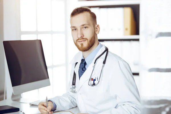 Vriendelijke rode baard dokter zittend en schrijvend aan klembord in zonnige kliniek. Geneesmiddelenconcept — Stockfoto