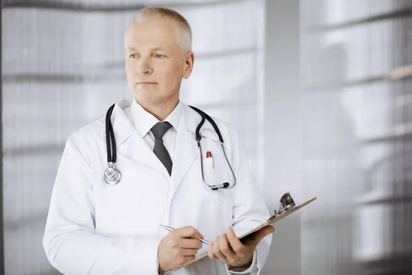 Elderly confident doctor standing straight and using clipboard in clinic. Perfect medical service. Medicine and healthcare concept