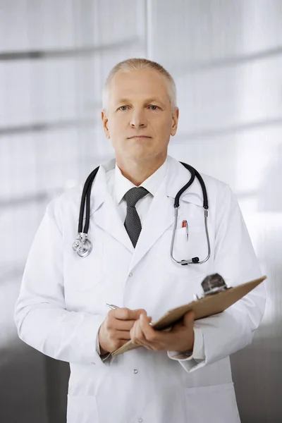 Ältere selbstbewusste Ärztin, die gerade steht und Klemmbrett in der Klinik benutzt. Perfekte medizinische Versorgung. Medizin und Gesundheitskonzept — Stockfoto