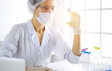 Koruyucu gözlüklü profesyonel kadın bilim adamı güneşli laboratuvarda reaktörlerle araştırma tüpü kullanıyor. Tıp ve araştırma.