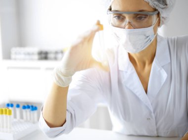 Koruyucu gözlüklü profesyonel kadın bilim adamı güneşli laboratuvarda reaktörlerle araştırma tüpü kullanıyor. Tıp ve araştırma.