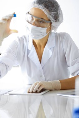 Koruyucu gözlüklü profesyonel kadın bilim adamı güneşli laboratuvarda reaktörlerle araştırma tüpü kullanıyor. Tıp ve araştırma.