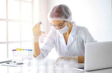 Koruyucu gözlüklü profesyonel kadın bilim adamı güneşli laboratuvarda reaktörlerle araştırma tüpü kullanıyor. Tıp ve araştırma.