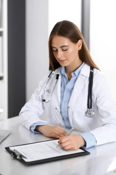 Läkarkvinnan fyller i läkarformuläret medan hon sitter vid skrivbordet på sjukhusets kontor. Läkare på jobbet. Medicin och hälsovård — Stockfoto