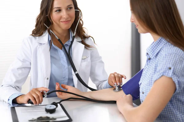 Az orvos ellenőrzi a női beteg vérnyomását, miközben az asztalnál ül a kórházi irodában. Kardiológia az orvostudományban és az egészségügyben — Stock Fotó