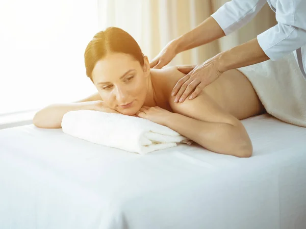 Ganska brunett kvinna njuter av proceduren med ryggmassage i solig spa salong. Skönhetskoncept. Kalltonad bild — Stockfoto