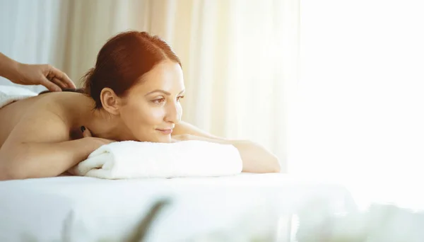 Mujer morena bonita disfrutando del tratamiento con piedras calientes en el salón de spa soleado. Concepto de belleza. Imagen tonificada en frío — Foto de Stock