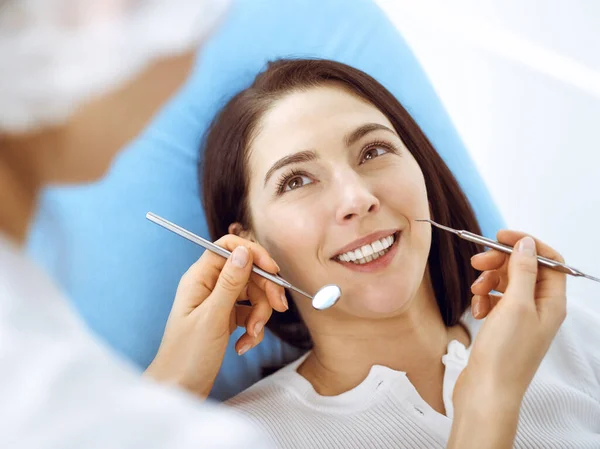 Lachende brunette vrouw wordt onderzocht door tandarts in de tandheelkundige kliniek. Handen van een arts met tandheelkundige instrumenten in de buurt van de mond van patiënten. Gezonde tanden en geneeskundeconcept — Stockfoto