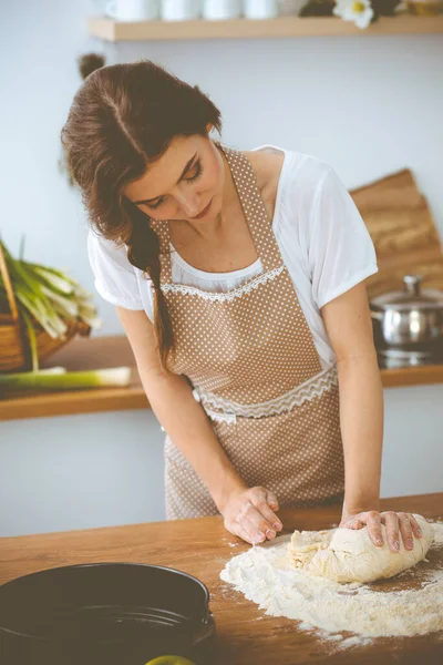 Jovem morena cozinhando pizza ou massa artesanal na cozinha. Dona de casa que prepara a massa de farinha na mesa de madeira. Conceito de alimentação, alimentação e saúde — Fotografia de Stock