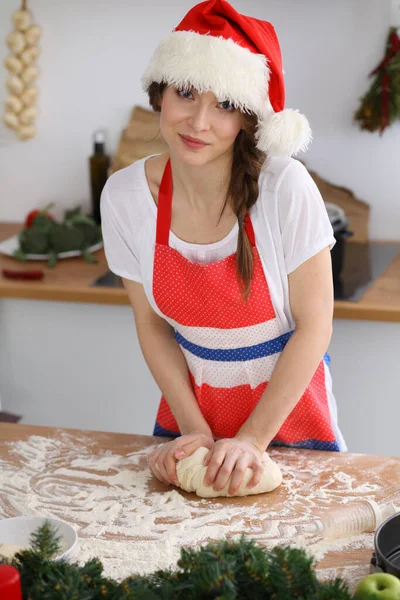 젊은 갈색 머리 여자 가 부엌에서 산타 클로스 모자를 쓰고 피자나 손으로 만든 파스타를 요리한다. 나무 식탁에서 반죽을 준비하는 주부 — 스톡 사진