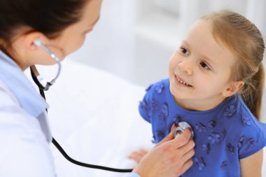 Doktor steteskopla küçük bir kızı muayene ediyor. Her zamanki tıbbi muayenede gülümseyen çocuk hasta. Tıp ve sağlık hizmetleri kavramları