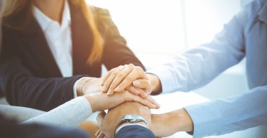 İş dünyası ekibi güneşli ofiste ellerini birleştiriyor. El ele tutuşan ve arkadaşlık, takım çalışması ve ortaklık kavramını temsil eden bir grup insan