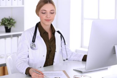 Hastanedeki genç bayan doktor masaüstü bilgisayar monitörüne bakıyor. Doktor ilaç geçmişi ve sınav sonuçlarını kontrol ediyor. Tıp ve sağlık hizmetleri kavramı