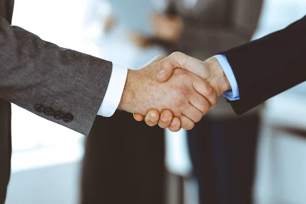 Business people shaking hands while standing with colleagues after meeting or negotiation, close-up. Group of unknown businessmen and women in modern office. Teamwork, partnership and handshake