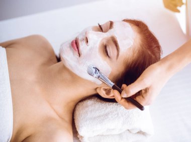Güzel esmer kadın güneşli spa merkezinde kapalı gözlerle kozmetik maske takmaktan zevk alıyor. Rahatlatıcı tedavi ve ilaç konsepti