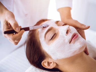 Güzel esmer kadın güneşli spa merkezinde kapalı gözlerle kozmetik maske takmaktan zevk alıyor. Rahatlatıcı tedavi ve ilaç konsepti