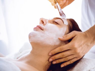 Güzel esmer kadın güneşli spa merkezinde kapalı gözlerle kozmetik maske takmaktan zevk alıyor. Rahatlatıcı tedavi ve ilaç konsepti