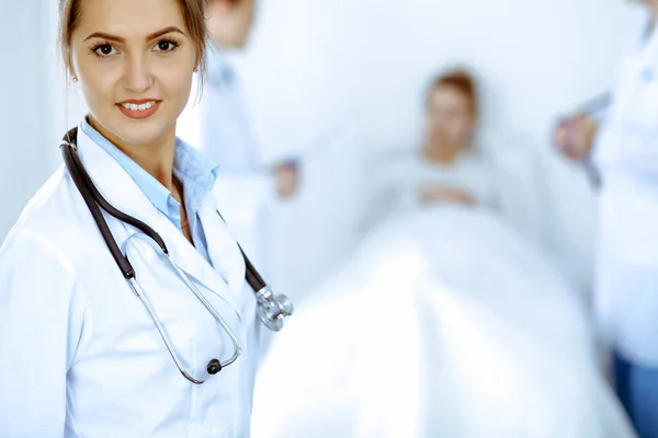 Жінка-лікар посміхається на фоні з пацієнтом в ліжку і двома лікарями — стокове фото