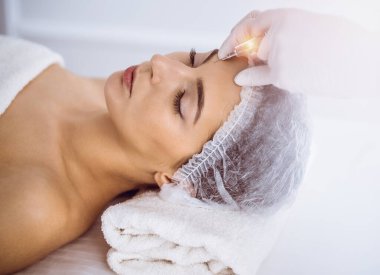 Güzellik uzmanı güneşli spa merkezinde genç esmer bir kadının yüzüne şırıngayla güzellik prosedürü uyguluyor. Kozmetik ilaç ve ameliyat, güzellik iğneleri.