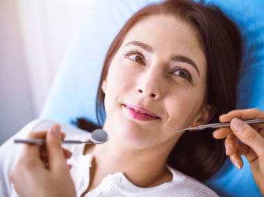 Güneşli diş kliniğinde dişçi tarafından muayene edilen gülümseyen esmer kadın. Hastanın ağzının yanında diş aletleri tutan bir doktorun elleri. Sağlıklı diş ve ilaç konsepti