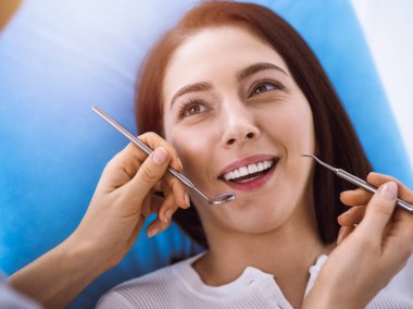 Güneşli diş kliniğinde dişçi tarafından muayene edilen gülümseyen esmer kadın. Hastanın ağzının yanında diş aletleri tutan bir doktorun elleri. Sağlıklı diş ve ilaç konsepti