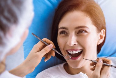 Güneşli diş kliniğinde dişçi tarafından muayene edilen gülümseyen esmer kadın. Hastanın ağzının yanında diş aletleri tutan bir doktorun elleri. Sağlıklı diş ve ilaç konsepti