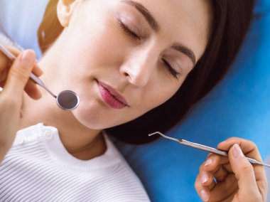 Güneşli diş kliniğinde dişçi tarafından muayene edilen gülümseyen esmer kadın. Hastanın ağzının yanında diş aletleri tutan bir doktorun elleri. Sağlıklı diş ve ilaç konsepti