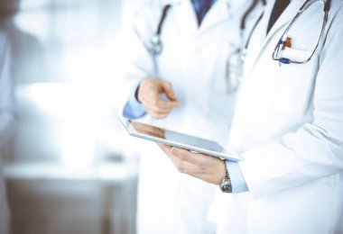 Bilinmeyen bir grup doktor hastane ofisinde dururken bazı tıbbi isim kayıtlarını kontrol etmek için bir bilgisayar tableti kullanıyor. Doktorlar hastaları muayene etmeye ve yardım etmeye hazır. Tıbbi yardım, sağlık sigortası, en iyi hastalık tedavisi ve ilaç dolandırıcılığı