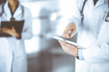 Bilinmeyen bir grup doktor hastane ofisinde dururken bazı tıbbi isim kayıtlarını kontrol etmek için bir bilgisayar tableti kullanıyor. Doktorlar hastaları muayene etmeye ve yardım etmeye hazır. Tıbbi yardım, sağlık sigortası, en iyi hastalık tedavisi ve ilaç dolandırıcılığı