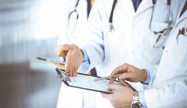 Steteskoplu bir grup doktor, erkek ve kadın hastane ofisinde ayakta dururken bir dosya ve tablet kullanarak tıbbi muayene sonuçlarını tartışıyor. Doktorlar ilaç kayıtlarını kontrol ediyor. Klinikte mükemmel bir sağlık hizmeti. 