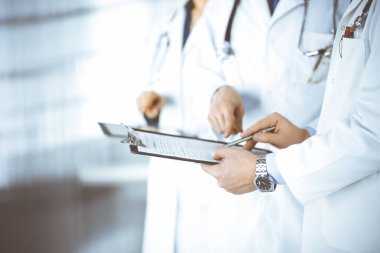 Steteskoplu bir grup doktor, erkek ve kadın hastane ofisinde ayakta dururken bir dosya ve tablet kullanarak tıbbi muayene sonuçlarını tartışıyor. Doktorlar ilaç kayıtlarını kontrol ediyor. Klinikte mükemmel bir sağlık hizmeti. 