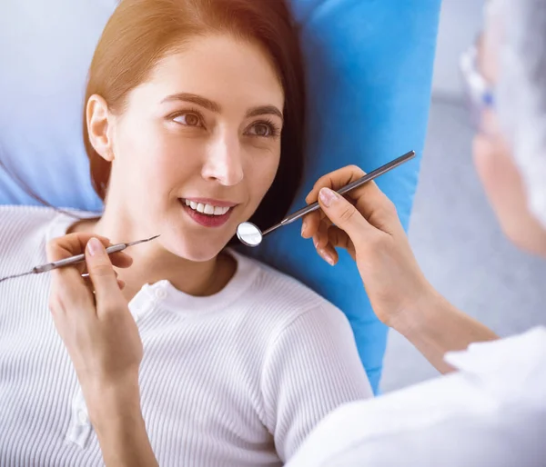 在阳光灿烂的牙科诊所接受牙医检查的黑发女人面带微笑。医生手拿着牙科器械靠近病人的嘴。健康的牙齿和医学概念 — 图库照片